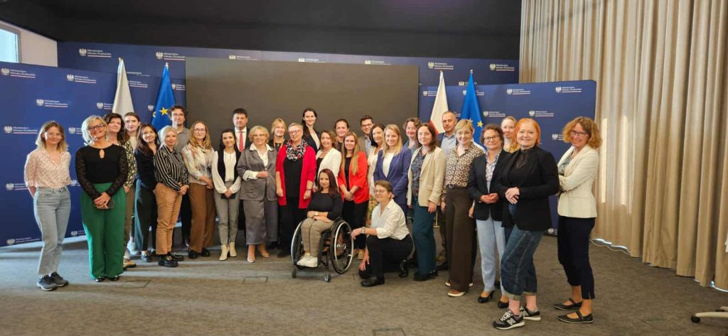 Zdjęcie grupowe uczestników spotkania w Ministerstwie Klimatu i Środowiska. Około 30 osób stoi i siedzi w kilku rzędach na tle ściany z logotypami ministerstwa oraz flagą Polski i flagą Unii Europejskiej. Wśród uczestników są zarówno kobiety, jak i mężczyźni, część osób uśmiecha się do obiektywu.