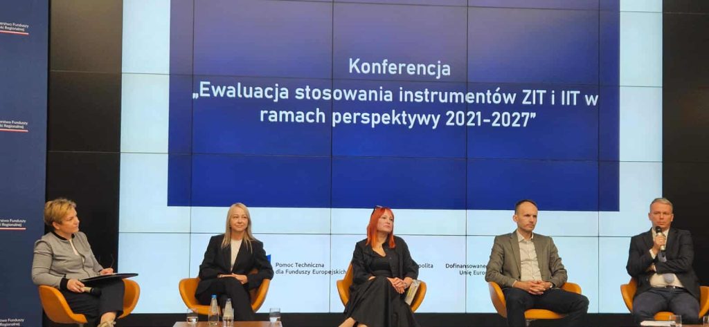 Pięć osób siedzi na scenie podczas konferencji pod tytułem „Ewaluacja stosowania instrumentów ZIT i IIT w ramach perspektywy 2021–2027”. W tle znajduje się duży ekran z tytułem wydarzenia. Uczestnicy panelu siedzą w żółtych fotelach, trzymają mikrofony i notatki. Na ekranie widoczne są także logotypy „Pomoc Techniczna dla Funduszy Europejskich” i „Dofinansowano ze środków Unii Europejskiej”.