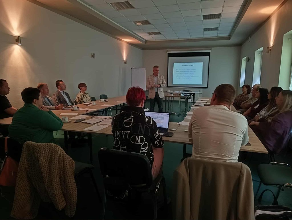 Na zdjęciu prowadzący szkolenie stoi przed grupą uczestników siedzących przy stołach ustawionych w podkowę. Na ekranie za nim wyświetlona jest prezentacja. W sali panuje robocza atmosfera, uczestnicy słuchają prelegenta i robią notatki.