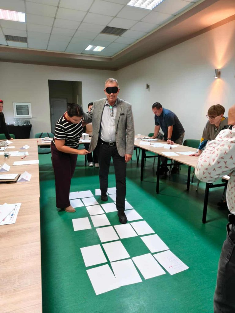 Na zdjęciu uczestnicy szkolenia biorą udział w ćwiczeniu polegającym na przeprowadzeniu osoby z zasłoniętymi oczami przez ścieżkę ułożoną z kartek papieru na podłodze. Pozostali uczestnicy obserwują i wspierają zadanie, które służy rozwijaniu zaufania i komunikacji w grupie.