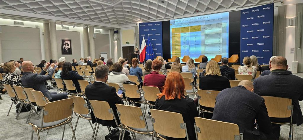 Uczestnicy konferencji siedzą na sali i słuchają prezentacji dotyczącej rewitalizacji. Na scenie, obok flag Polski i Unii Europejskiej, stoi prelegentka przemawiająca przy mównicy. Za nią znajduje się duży ekran z tabelą prezentującą dane oraz rząd pustych żółtych foteli dla panelistów. W tle widoczna jest niebieska ścianka z logo Ministerstwa Funduszy i Polityki Regionalnej.