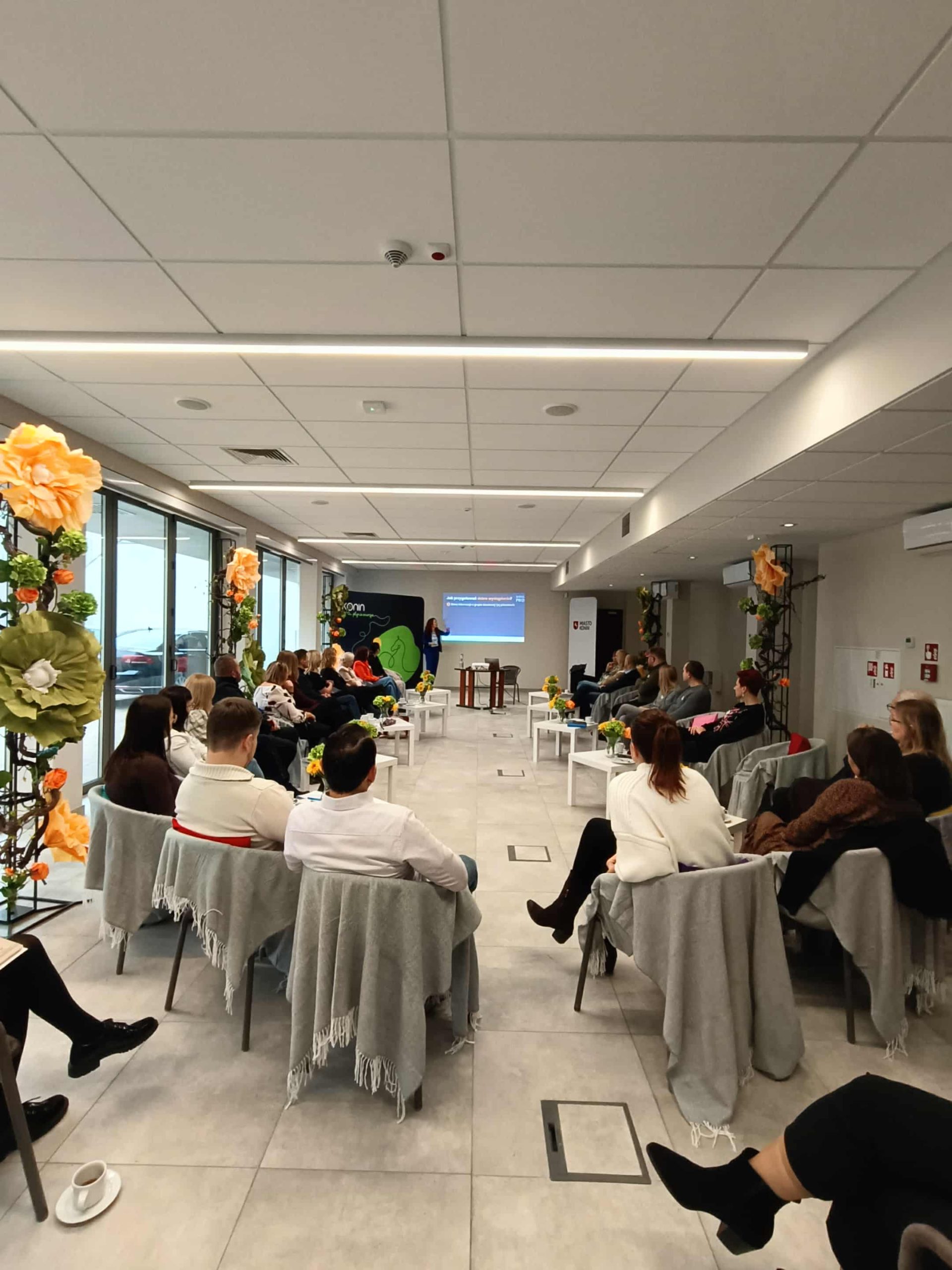 Grupa uczestników szkolenia siedzi w półokręgu w jasnej sali z dużymi oknami. Na środku ustawiono małe stoliki z notatkami, filiżankami i wazonami z kwiatami. Przed grupą stoi prowadzący prezentację mężczyzna, a za nim wyświetlany jest slajd. Wnętrze udekorowane jest kwiatami, a uczestnicy uważnie słuchają i prowadzą notatki.