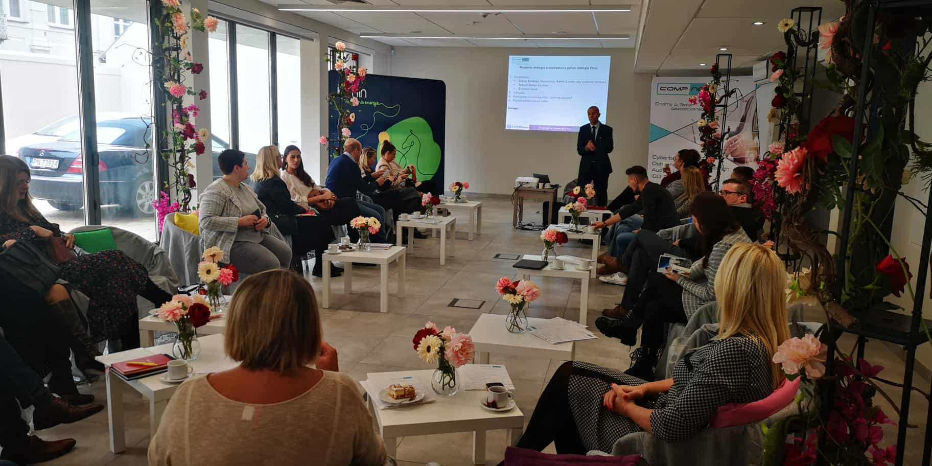 Grupa uczestników szkolenia siedzi w półokręgu w jasnej sali z dużymi oknami. Na środku ustawiono małe stoliki z notatkami, filiżankami i wazonami z kwiatami. Przed grupą stoi prowadzący prezentację mężczyzna, a za nim wyświetlany jest slajd. Wnętrze udekorowane jest kwiatami, a uczestnicy uważnie słuchają i prowadzą notatki.