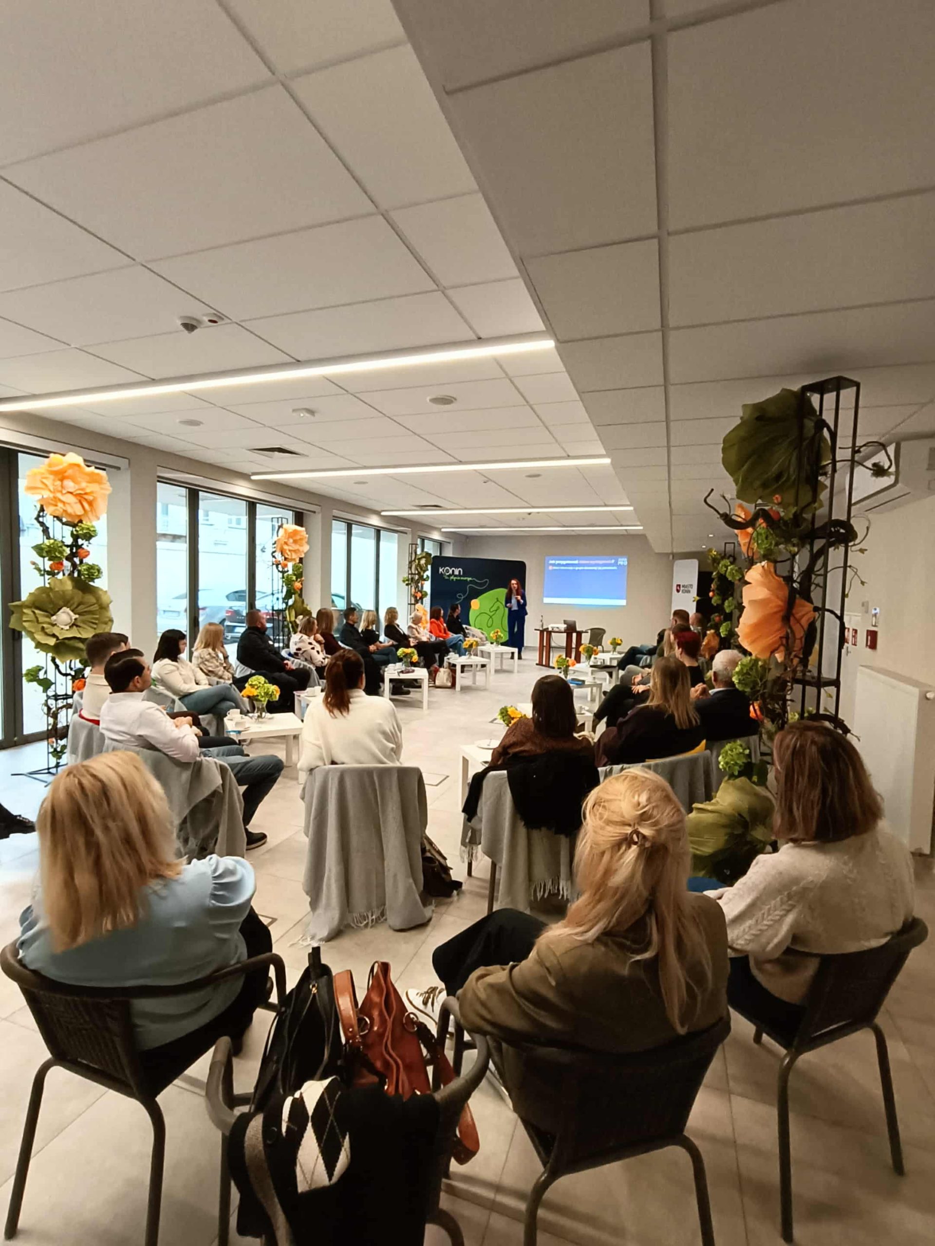 Grupa uczestników szkolenia siedzi w półokręgu w jasnej sali z dużymi oknami. Na środku ustawiono małe stoliki z notatkami, filiżankami i wazonami z kwiatami. Przed grupą stoi prowadzący prezentację mężczyzna, a za nim wyświetlany jest slajd. Wnętrze udekorowane jest kwiatami, a uczestnicy uważnie słuchają i prowadzą notatki.