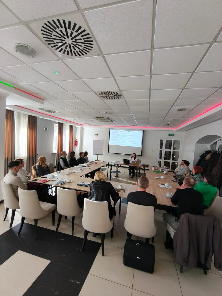 Fotografia przedstawia grupę osób siedzących przy dużym stole w kształcie litery U w jasnej sali konferencyjnej. Na czele stołu stoi prowadząca szkolenie, Maria Michułka, a obok niej znajduje się ekran z wyświetloną prezentacją na temat zamówień publicznych. Uczestnicy szkolenia siedzą na nowoczesnych, jasnych krzesłach, słuchając wykładu. Na stołach widoczne są filiżanki z kawą i materiały szkoleniowe.