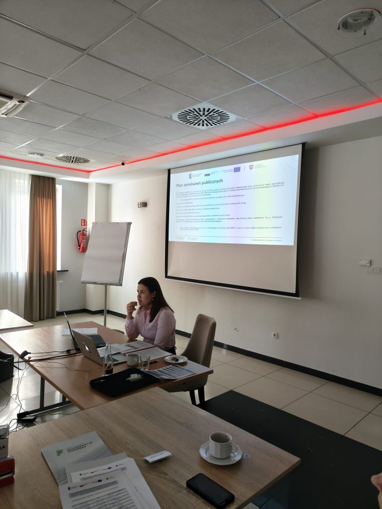 Zdjęcie portretowe Marii Michułki, uśmiechniętej kobiety o spiętych włosach, ubranej w białą koszulę i ciemną marynarkę. W tle widoczne są logotypy Funduszy Europejskich, Rzeczypospolitej Polskiej oraz Samorządu Województwa Wielkopolskiego.