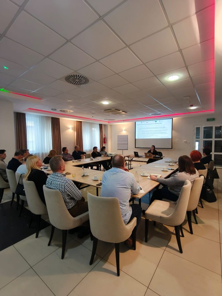 Fotografia przedstawia grupę osób siedzących przy dużym stole w kształcie litery U w jasnej sali konferencyjnej. Na czele stołu stoi prowadząca szkolenie, Maria Michułka, a obok niej znajduje się ekran z wyświetloną prezentacją na temat zamówień publicznych. Uczestnicy szkolenia siedzą na nowoczesnych, jasnych krzesłach, słuchając wykładu. Na stołach widoczne są filiżanki z kawą i materiały szkoleniowe.