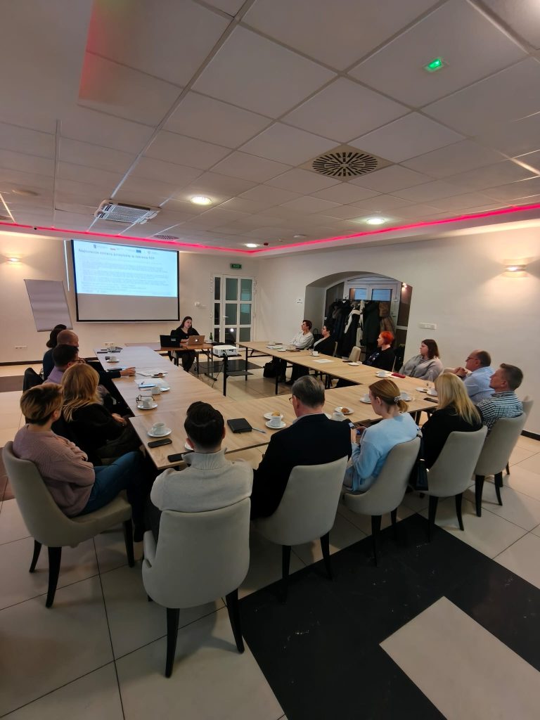 Fotografia przedstawia grupę osób siedzących przy dużym stole w kształcie litery U w jasnej sali konferencyjnej. Na czele stołu stoi prowadząca szkolenie, Maria Michułka, a obok niej znajduje się ekran z wyświetloną prezentacją na temat zamówień publicznych. Uczestnicy szkolenia siedzą na nowoczesnych, jasnych krzesłach, słuchając wykładu. Na stołach widoczne są filiżanki z kawą i materiały szkoleniowe.